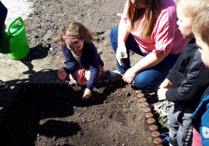 Zdjęcie przedstawia dziewczynkę wraz z nauczycielką sadzącą nasiona oraz dzieci obserwujące czynność sadzenia